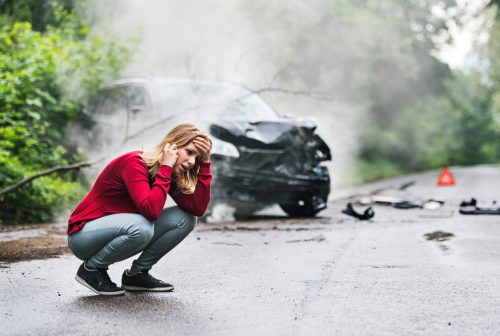 A young woman with smartphone by the damaged car after a car accident, making a phone call