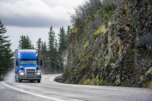 Big rig blue semi truck with grill guard transporting cargo in semi trailer running on the winding wet road with rain dust with rock cliff on the side