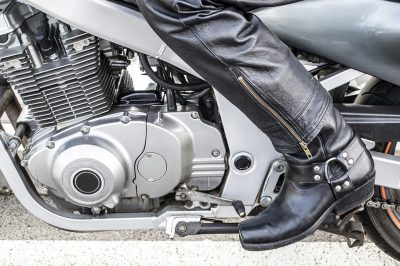 Leg of a man in black leather pants and boots standing in front of chrome motorcycle frame and engine