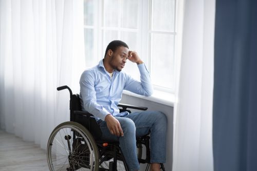 Unhappy black handicapped guy in wheelchair looking out window, feeling sad and desperate at home, copy space