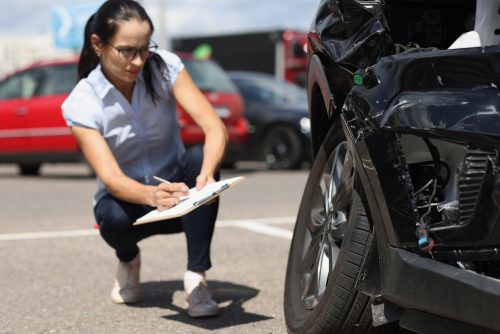 Woman inspector writing in documents on clipboard near broken car. Auto insurance concept