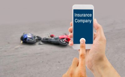 hand holding a cellphone with insurance company on the screen with a motorcycle accident in background